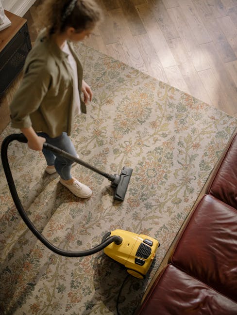 A person using a yellow and black vacuum cleaner on a patterned carpet in a residential room, with their hands holding a cloth to clean or adjust the vacuum's dust container. The carpet appears clean