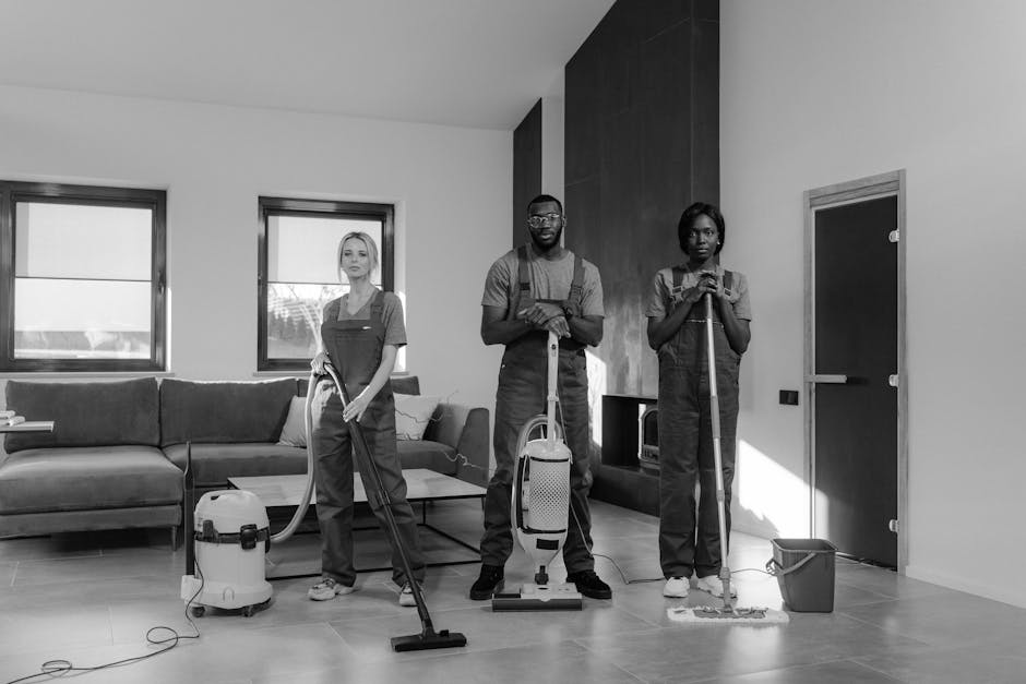 Three professional cleaners from Cleaners Shepherds Bush are positioned in a modern living room, performing surface cleaning and deep cleaning tasks. The room features a tiled floor, a dark-colored se